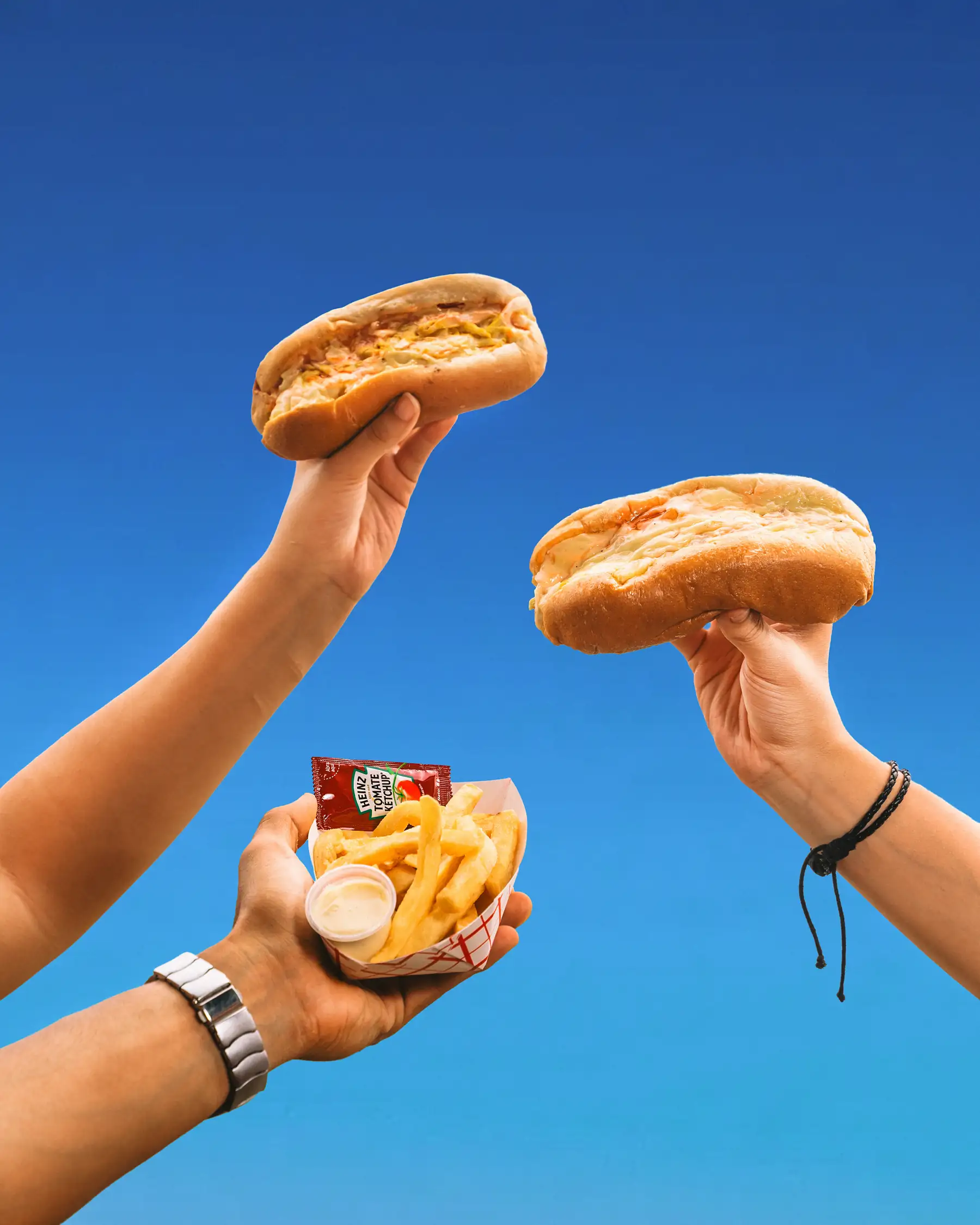 Dos manos sostienen hot dogs Marusha's contra un cielo azul, con una charola de papas al fondo